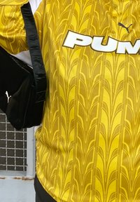 Geltonas raštuotas sportinis marškinėliai su PUMA logotipu. Juoda petneša krepšys pirmame plane. Medžiaga atrodo lengva ir pralaidi.