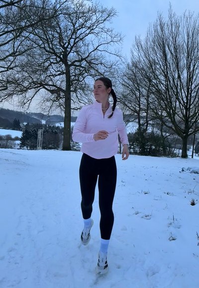 Mujer con chaqueta rosa y leggings negros caminando por un camino nevado rodeado de árboles desnudos en un paisaje invernal.