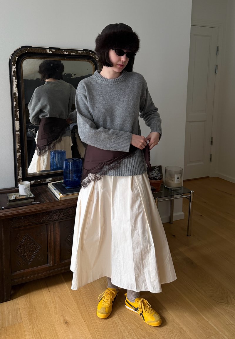 Femme portant un pull gris, une jupe crème, une écharpe marron, des baskets jaunes et des lunettes de soleil foncées, debout près d'un miroir et d'une armoire en bois à l'intérieur.