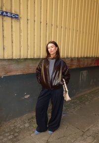 Chaqueta bomber marrón y brillante sobre una camisa a rayas en blanco y negro, combinada con pantalones negros holgados y calcetines azul claro. Sosteniendo un bolso pequeño.