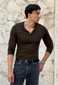 Hnědá dlouhá rukávová henley košile s knoflíkovou légou, doplněná tmavými džíny. Outfit obsahuje černý kožený opasek a stříbrné hodinky.