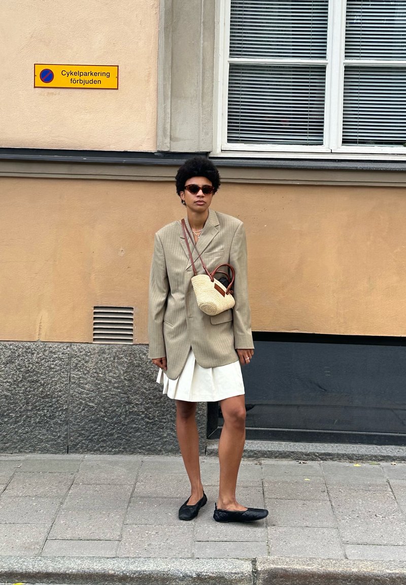 Μπεζ ριγέ oversized σακάκι, λευκή φούστα, μαύρες μπαλαρίνες και υφασμένη τσάντα. Το μοντέλο φοράει γυαλιά ηλίου και στέκεται μπροστά σε μια υφασμένη τοίχο.