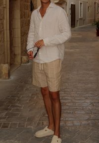 Svetla bež lanena srajca dolgih rokavov, v kombinaciji z bež črtastimi kratkimi hlačami in belimi natikači. Nastavitev vsebuje tlakovane ulice.
