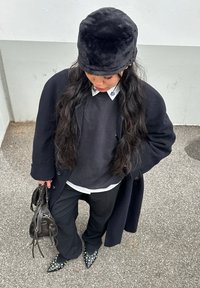 Zwarte nepbontemmerhoed, zwarte jas, grijze sweatshirt over een witte blouse, zwarte broek, gestructureerde puntige hakken, met een zwarte handtas.