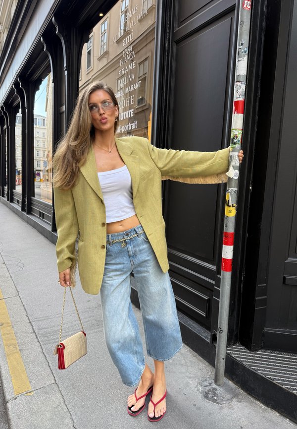 Femme aux longs cheveux portant un blazer jaune, un crop top blanc, un jean large et des sandales rouges, tenant un poteau et un petit sac à main sur un trottoir en ville.