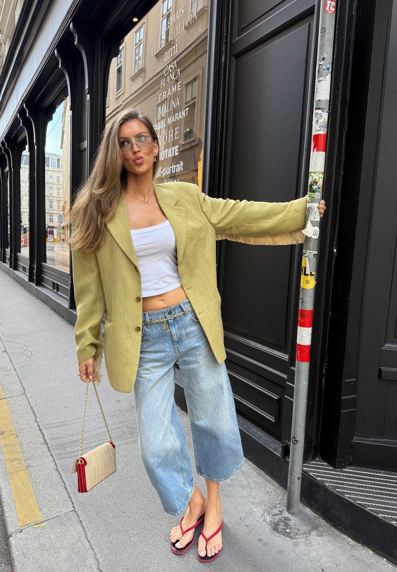 Femme aux longs cheveux portant un blazer jaune, un crop top blanc, un jean large et des sandales rouges, tenant un poteau et un petit sac à main sur un trottoir en ville.
