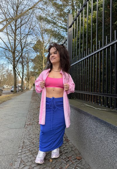 Mujer de pie en una acera cerca de una cerca negra, vestida con un top corto rosa, camisa rosa, falda azul y zapatillas blancas, con árboles de fondo.