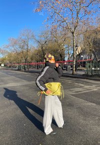 Personne portant une veste noire, un pantalon blanc, un chapeau à motifs et des lunettes de soleil, tenant un sac jaune tout en se tenant dans une rue urbaine ensoleillée.