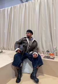 Chaqueta marrón forrada de piel de oveja con detalles en negro, suéter gris, pantalones cortos anchos de mezclilla y botas negras de tacón. Ambientado contra un fondo beige.