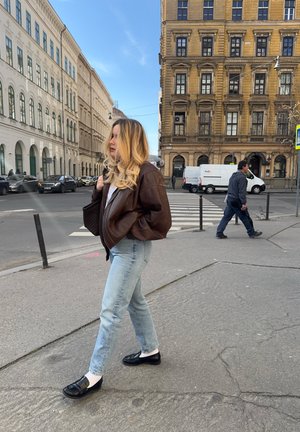 Mujer con cabello rubio, vestida con chaqueta marrón y jeans, camina por la acera de la ciudad cerca de un paso de peatones y edificios históricos en un día despejado.