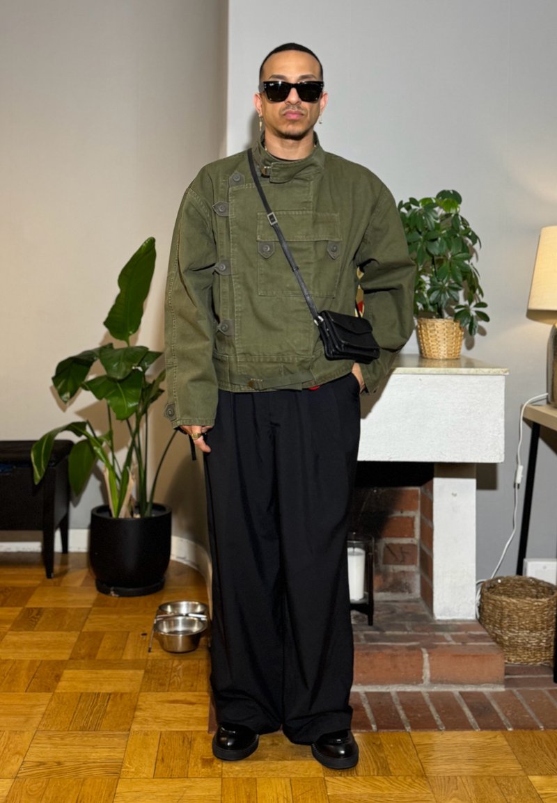 Man wearing dark sunglasses, olive green jacket, black wide trousers, and crossbody bag stands indoors near plants and a fireplace.