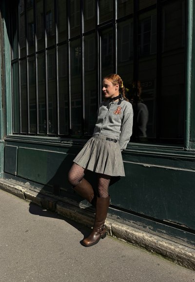 Cárdigan gris con bordado de oso de peluche, falda de cuadros negros y blancos con plisados, medias de rejilla y botas marrones hasta la rodilla contra una pared verde.