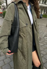 Cappotto lungo trapuntato verde oliva con due bottoni e una borsa a tracolla in pelle nera testurizzata. Il modello indossa una camicia bianca e un giubbotto di jeans sotto.