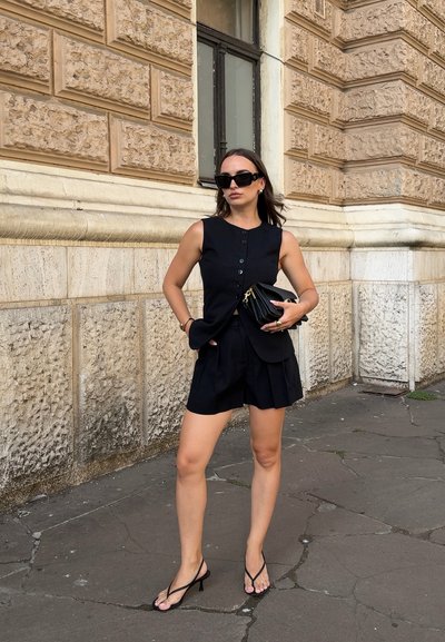 Blusa negra sin mangas con botones en la parte delantera, combinada con pantalones cortos negros con pliegues y sandalias de tacón con tiras. Un bolso de mano negro y unas gafas de sol grandes completan el look.
