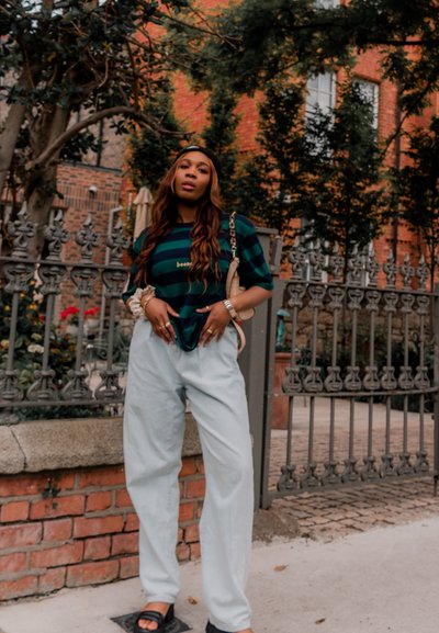 Camiseta a rayas verde y azul marino con texto, pantalones relajados azul claro, sandalias negras y un bolso beige. Pared de ladrillo y vegetación al fondo.