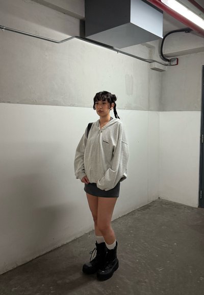 Joven mujer con cabello negro en trenzas, vestida con una sudadera gris con cremallera, una mini falda oscura, calcetines blancos y botas de plataforma negras, de pie en una sala de concreto.