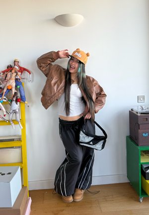 Joven con atuendo informal posando en interiores, lleva una chaqueta de cuero marrón, gorra naranja con orejas, pantalones negros y sostiene una bolsa pequeña.