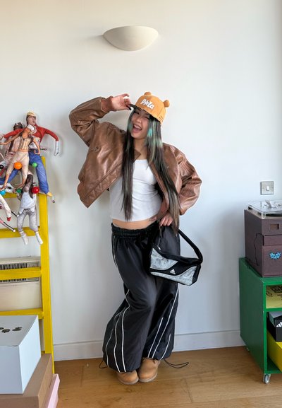 Joven con atuendo informal posando en interiores, lleva una chaqueta de cuero marrón, gorra naranja con orejas, pantalones negros y sostiene una bolsa pequeña.