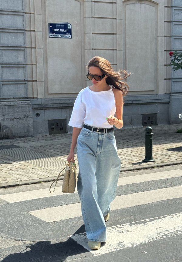 Kvinde med langt hår, solbriller, hvid asymmetrisk top og brede jeans, der går over et fodgængerfelt med en vævet håndtaske.