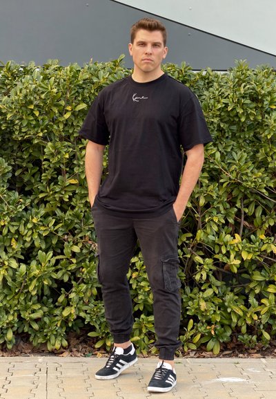 Hombre con camiseta negra, pantalones cargo oscuros y zapatillas negras y blancas, de pie con las manos en los bolsillos frente a arbustos verdes.