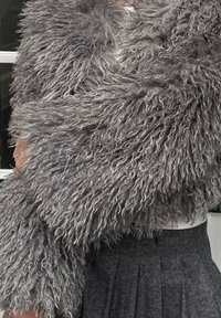 Umelý kožušinový plášť v šedej farbe s dlhou, kučeravou textúrou; mäkký vzhľad. Dopĺňa ho štruktúrovaná sivá sukňa. Biela blúzka pod ním je čiastočne viditeľná.