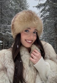 Jeune femme souriante dans une forêt enneigée portant un grand chapeau en fourrure beige et un manteau assorti en fourrure, tenant le col du manteau d'une main.