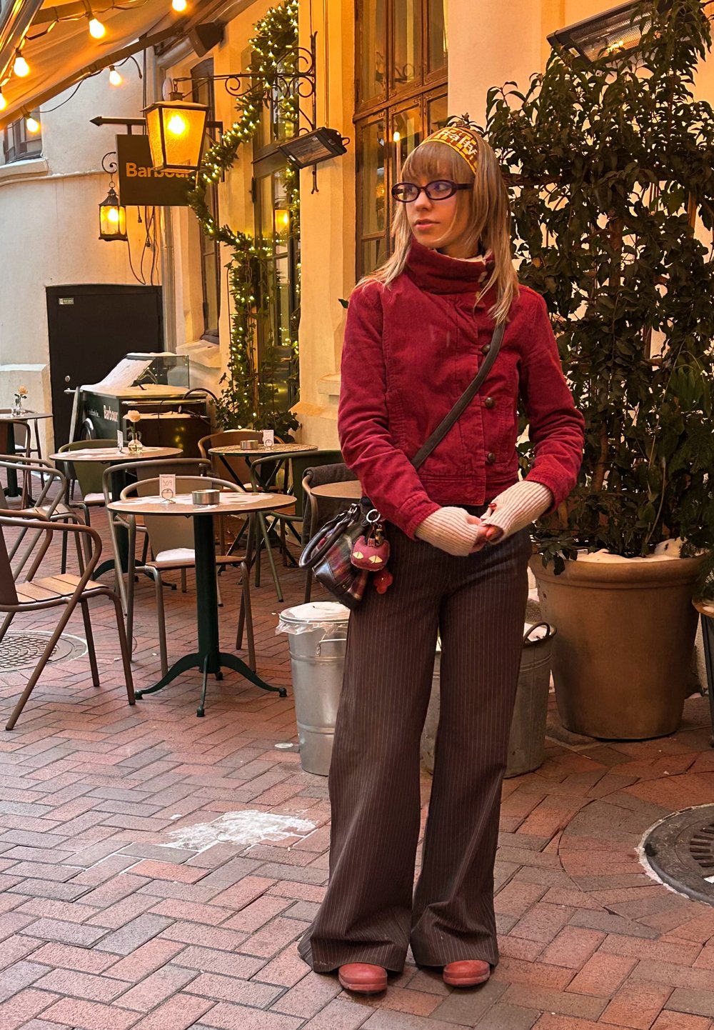 Veste en velours côtelé rouge avec un col montant, associée à un pantalon large marron à fines rayures et des sabots en bois. Comprend des gants gris et un petit sac.