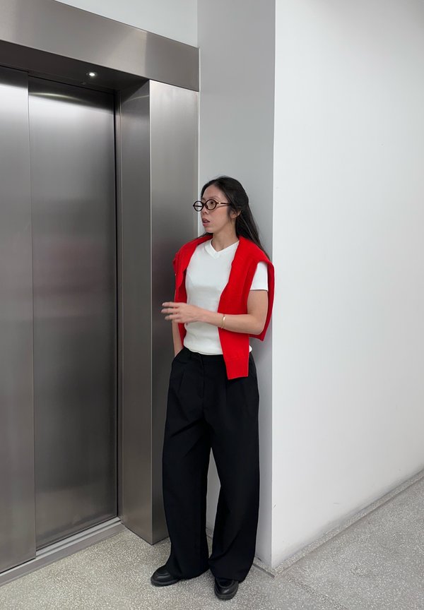 Una mujer con gafas y un jersey rojo espera junto a las puertas cerradas de un ascensor en un pasillo moderno.