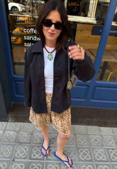 Mujer con gafas de sol, chaqueta negra, camiseta blanca, falda de lunares, chanclas azules y collar, de pie en la acera junto a una cafetería.