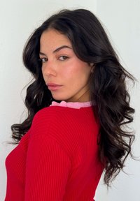 Rød ribstrikket sweater med lyserød krave, med langt mørkt bølget hår, lys hud med fregner og set i profil.