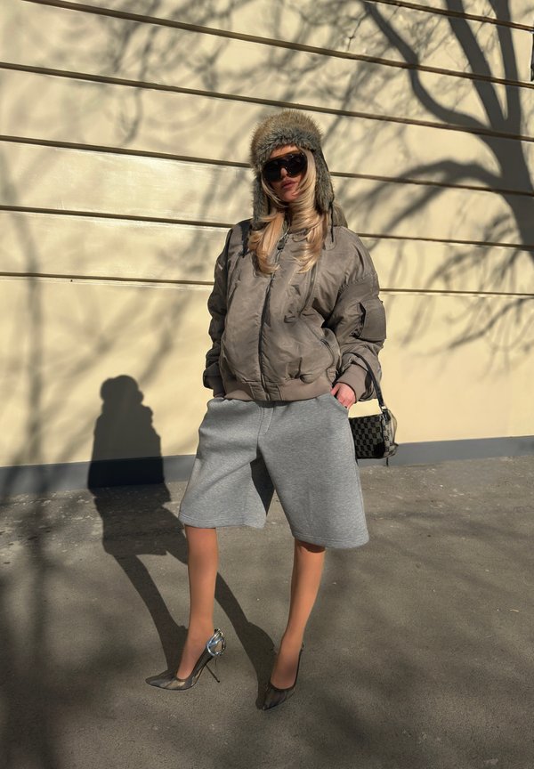 Femme portant une veste bomber surdimensionnée, un short large gris, des talons hauts, un chapeau de trappeur en fourrure et des lunettes de soleil, debout contre un mur avec des ombres d'arbres.