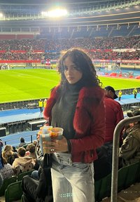 Crvena jakna s krznenim akcentima, crni rolak, svijetloplave traperice. Drži šarene šalice na sportskom događaju, s prepunim stadionom u pozadini.