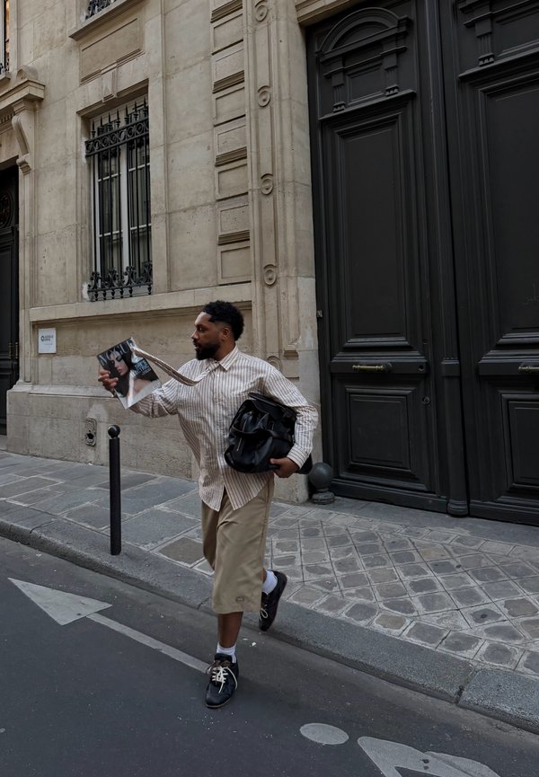Homme marchant dans une rue de la ville, tenant un magazine de mode dans une main et un sac noir dans l’autre, passant devant un bâtiment avec de grandes portes.