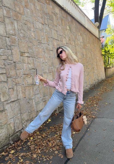 Cárdigan rosa claro con acentos de volantes, blusa blanca, jeans acampanados azules claros y botas de tobillo marrones, sosteniendo una bebida y un bolso marrón.