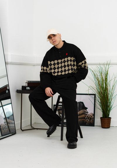 Suéter de polo negro con un patrón de diamantes en tono beige, logo rojo, pantalones negros y zapatos negros. Gorra blanca y un ambiente interior minimalista.