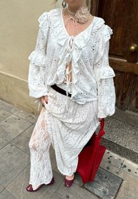 Fehér csipkeblúz fodros ujjakkal, csipkés szoknya virágmintás anyag fölé rétegezve. Piros táska és bordó, lekerekített orrú cipő.