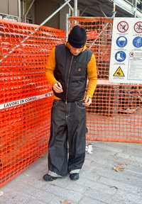 Melnā pūkainā veste pār oranžu kapuci džemperi, vaļīgas tumši zila džinsa bikses un melni apavi. Fonā redzams oranžs būvniecības siets un norādes.