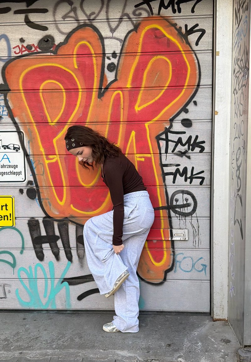 Jeune femme en haut marron et pantalon de survêtement gris ajustant sa chaussure devant un graffiti coloré avec de grandes lettres "PWR" sur le mur.