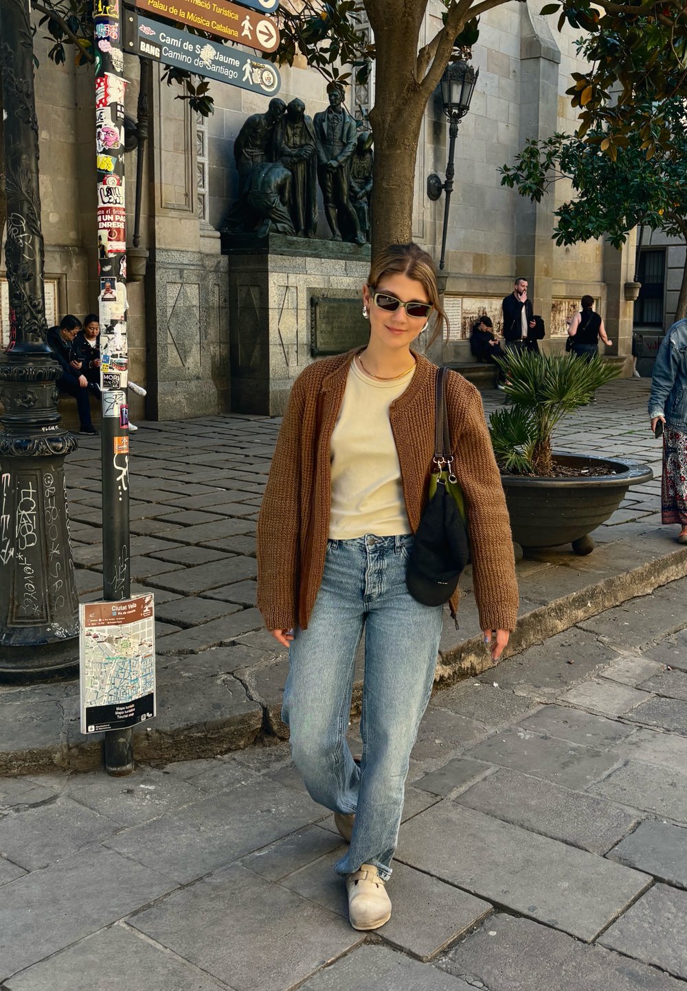 Cardigã castanho de malha sobre uma t-shirt clara, jeans azuis e sapatos bejes. Acessórios incluem óculos de sol e uma mala preta. Fundo de rua de calçada.