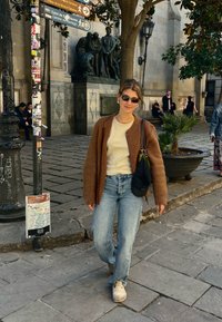 Cardigã castanho de malha sobre uma t-shirt clara, jeans azuis e sapatos bejes. Acessórios incluem óculos de sol e uma mala preta. Fundo de rua de calçada.