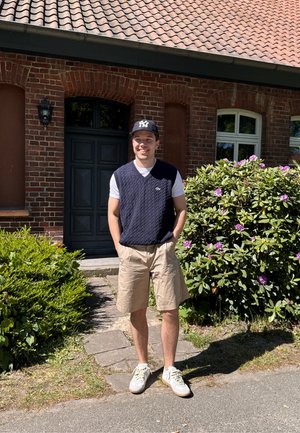 Hombre de pie frente a una casa de ladrillo, vistiendo un chaleco de punto azul marino sobre una camiseta blanca, pantalones cortos beige y zapatillas claras. Hay follaje verde y flores cerca.