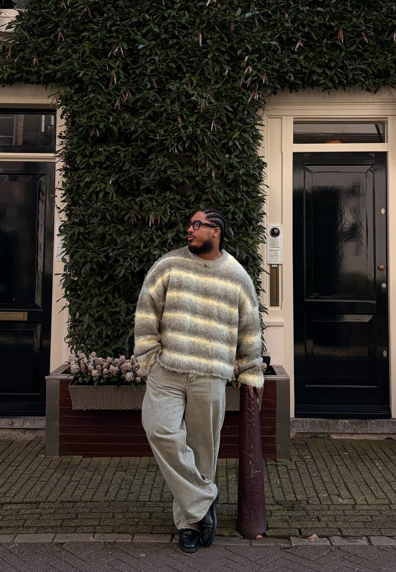 Homme aux cheveux tressés et lunettes portant un pull rayé et duveteux ainsi qu'un pantalon clair, appuyé sur un poteau devant un mur couvert de vignes et des portes noires.