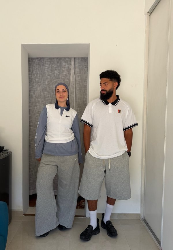 Deux personnes sont debout à l'intérieur contre un mur blanc ; l'une porte un foulard, un gilet blanc et un pantalon gris ample, l'autre porte un polo blanc et un short gris.