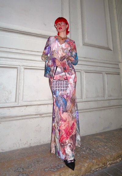 Vestido largo y multicolor con patrones abstractos de remolinos en tonos de rosa, púrpura y dorado. Llevado con un pequeño bolso texturizado y tacones puntiagudos.