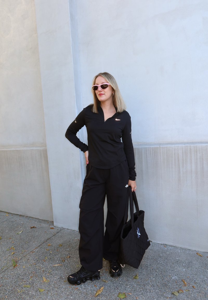 Jeune femme en tenue de sport noire Nike et lunettes de soleil se tient contre un mur gris clair, tenant un sac fourre-tout matelassé noir avec un logo blanc.