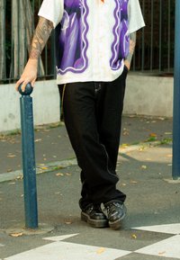 Short-sleeved white shirt with a purple graphic design, black trousers, and black platform shoes, standing next to a blue pole on the pavement.
