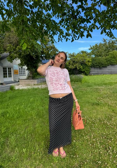Camiseta rosa claro con estampados de dibujos animados, falda maxi de lunares negros, chanclas rosas y un bolso de mimbre naranja. Fondo de hierba verde.