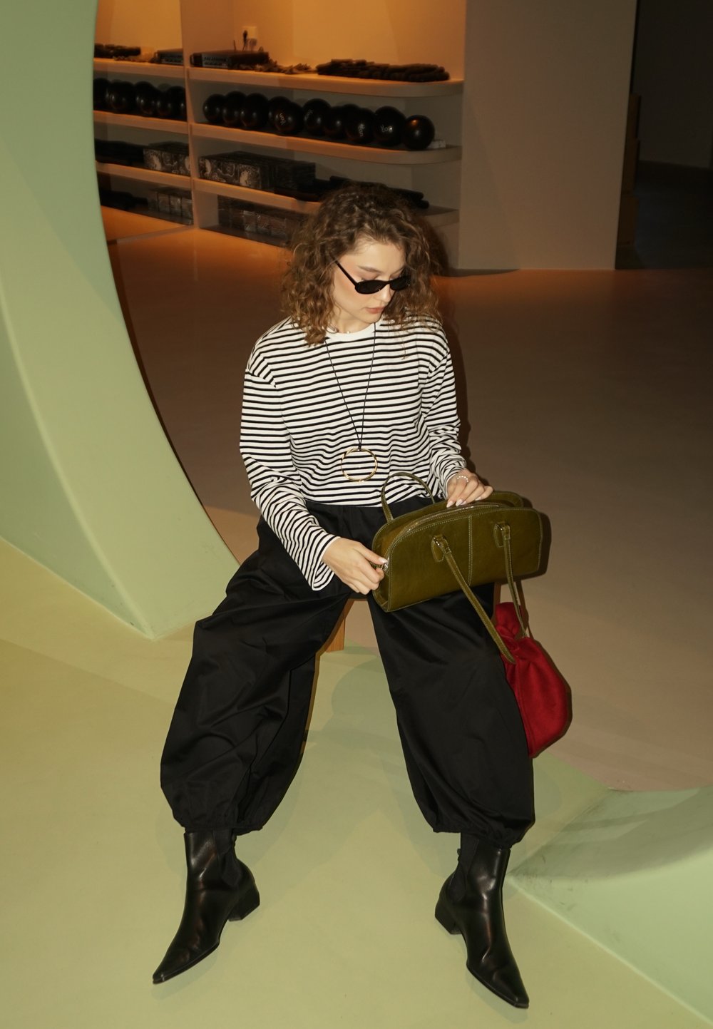Haut à manches longues rayé, pantalon noir oversized, bottines noires ; tenant un sac à main vert olive avec deux anses et un sac rouge à côté.