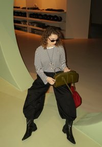 Haut à manches longues rayé, pantalon noir oversized, bottines noires ; tenant un sac à main vert olive avec deux anses et un sac rouge à côté.