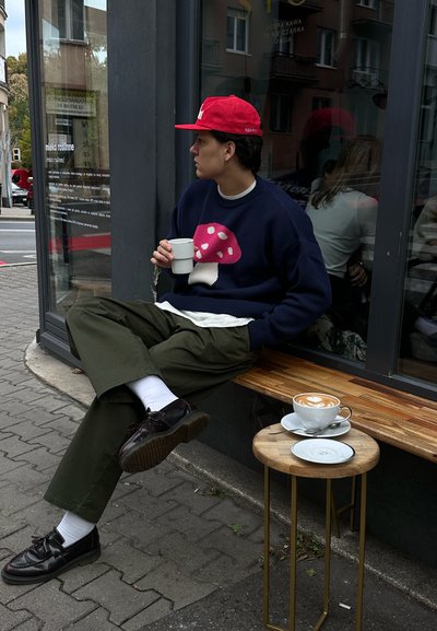 Suéter azul marino con un diseño de hongo rosa, pantalones oliva, calcetines blancos y zapatos negros; sosteniendo una taza blanca. Café en un taburete de madera.